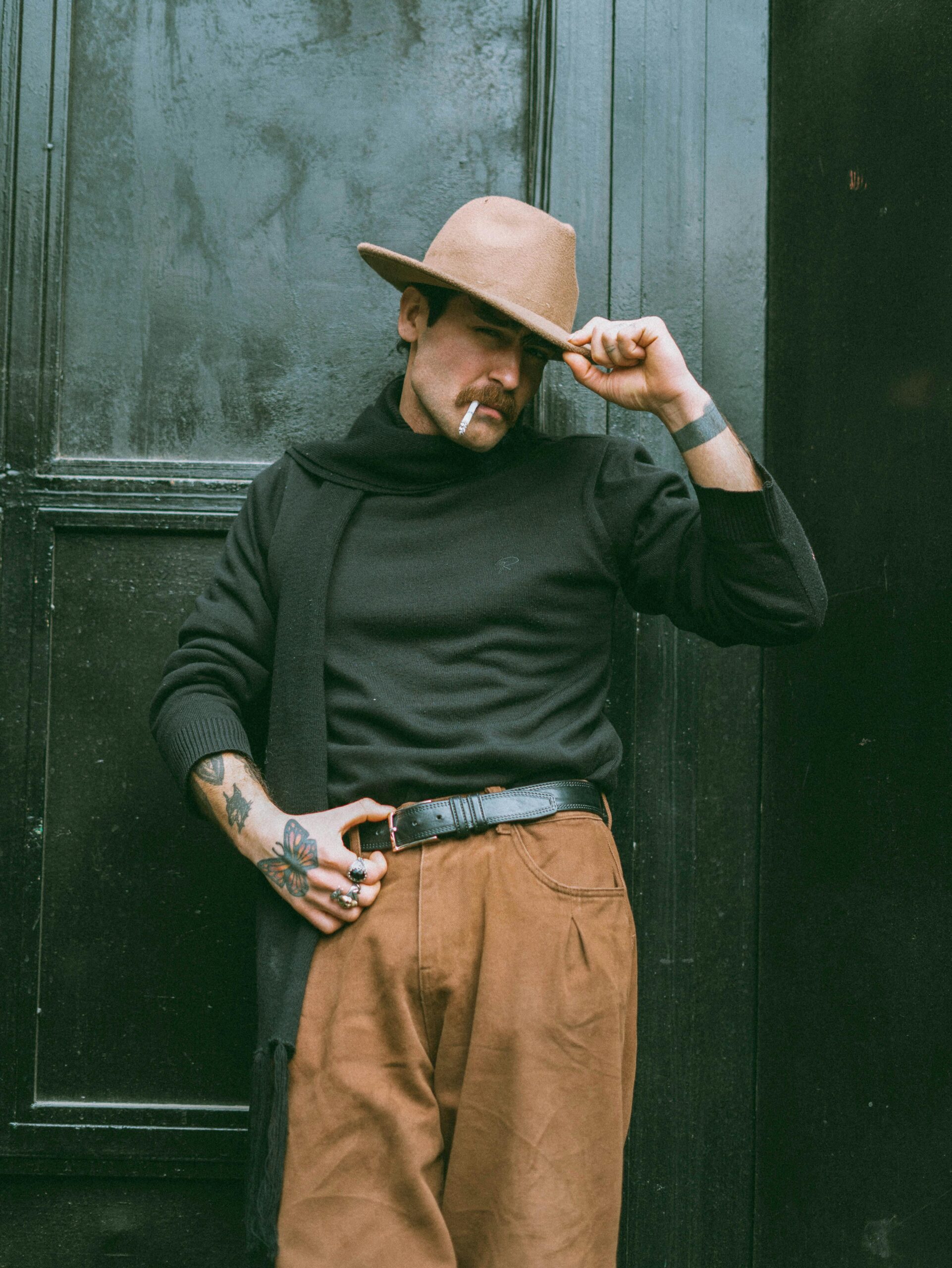 Fashionable man with tattoos and a hat posing confidently against a black door.